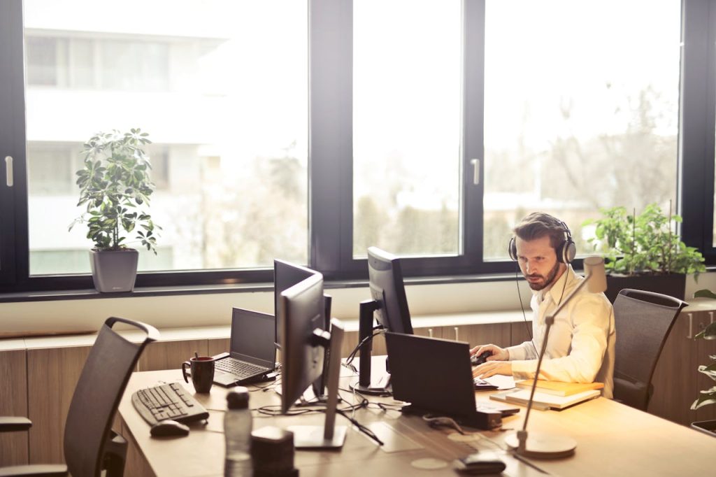 man-with-headphones-facing-computer-monitor-845451 A businessman sits at a desk using multiple computers and a headset in a well-lit modern office.